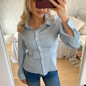 Åtsittande skjorta  - Säljer en stilren blå- och vitrandig skjorta med långa ärmar och knappar framtill. Skjortan har lite tajtaste passform och passar storlek S-M. Knappt använd. 