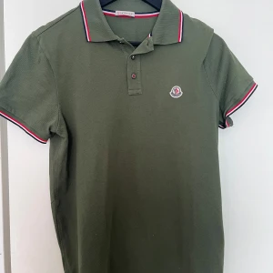 Grön pikétröja från Moncler - Snygg grön pikétröja från Moncler med klassisk krage och knappar. Tröjan har korta ärmar med röd och vit randdetalj på krage och ärmslut. Perfekt för en stilren look.