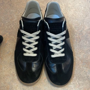 Svarta sneakers från Maison Margiela - Snygga svarta sneakers från Maison Margiela med vit snörning och en stilren design. Skorna har en kombination av mocka och skinn, vilket ger en lyxig känsla. Perfekta för dig som vill ha en stilren look. Box är slängd ‼️, pris kan diskuteras vid snabb affär.  Använda cirka en vecka 