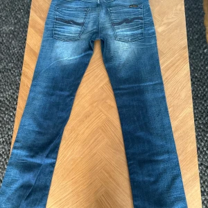 Nudie jeans med feta slitningar  - Snygga blå jeans från Nudie Jeans med en klassisk femficksdesign och dragkedja. Jeansen har en lätt tvättad look med subtila slitningar för en trendig stil. Perfekta för en avslappnad vardagslook. Storleken är W31 L32 och är perfekt och väldigt trendiga för tillfället. Hör av dig ifall du har några frågor!