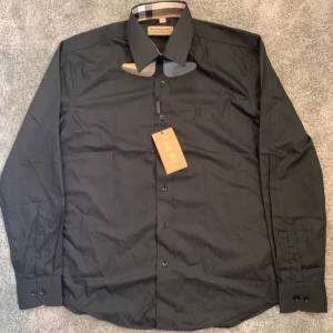 Svart skjorta från Burberry - Elegant svart skjorta från Burberry med klassisk krage och långa ärmar. Skjortan har en diskret logotyp broderad på bröstet och knappar framtill. Perfekt för en stilren look.