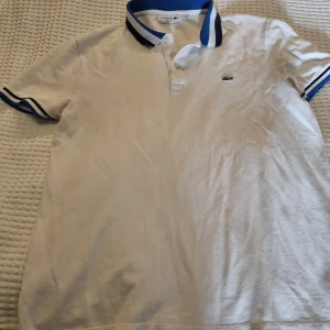 Vit pikétröja från Lacoste - Vit pikétröja från Lacoste med blå och svarta detaljer på kragen och ärmsluten. Tröjan har en broderad logotyp på bröstet och knappar framtill.