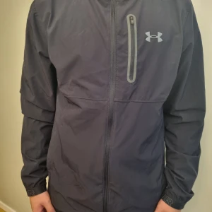 Under Armour jacka - Svart vindjacka från Under Armour. Jackan har en dragkedja framtill och en praktisk bröstficka med dragkedja.