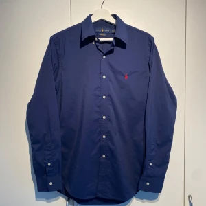 Ralph Lauren Performance Navy Skjorta - Ralph Lauren skjorta i marinblå färg. Performance material vilket gör den mycket mer bekväm och en mer matt yta. Använd en del, okej skick. Små fläckar men går bort i tvätten. Den har en broderad röd logotyp på bröstet som ger en stilren touch. Stilsäker, funkar lika bra i vardagen som på krogen. Nypris 1700kr. Storlek M och true to Size. Fraktas inom ett dygn. Skriv vid frågor eller funderingar. 