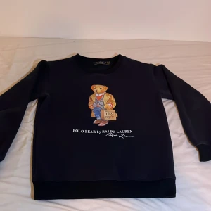 Svart sweatshirt med Polo Bear från Ralph Lauren - Säljer en svart sweatshirt från Ralph Lauren med det ikoniska Polo Bear-motivet på framsidan. Tröjan har långa ärmar och en rund halsringning. Perfekt för en avslappnad stil med en touch av lyx.