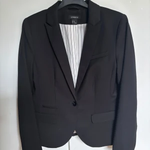 Svart kavaj från Lindex - Snygg svart kavaj från Lindex med en klassisk design. Toppskick. Perfekt använda som jacka eller som finkavaj till kjol. Fint material, elastan, polyester och viskos. Normal passform.
