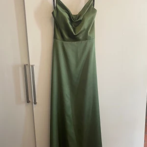 Grön långklänning i satin - Elegant grön långklänning i satin med smala axelband och en vacker drapering framtill. Klänningen har en öppen rygg med knytband för en justerbar passform. Klänningen är i storlek 38 men kan passa 36/40 eftersom man enkelt kan justera med snörena i ryggen. Kommer med en fin sjal. Passar till tex. bal och bröllop. Köpt på Johans högtidskläder. Kvitto finns.  Nypris: 3200kr Inköpt 2024