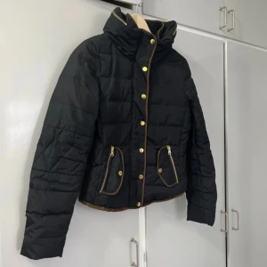 Svart quiltad jacka med dragkedja och knappar Small - Vero Moda, svart quiltad jacka med både dragkedja och knappar framtill. Kort modell med huva som går att dölja med dragkedja. Färgskav finns på knappar enligt bild.