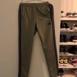 Gröna träningsbyxor från Adidas - Snygga gröna träningsbyxor från Adidas med svarta ränder längs sidorna. Byxorna har en elastisk midja och två sidofickor. Perfekta för träning eller en avslappnad stil. Byxorna är i storlek S. Byxorna är i mycket bra skick. 