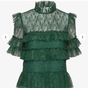 by malina blus - Elegant grön spetsblus med volanger och hög krage. Perfekt för en stilfull look med sina genomskinliga detaljer och korta ärmar. Blusen har en romantisk känsla och passar utmärkt för speciella tillfällen.