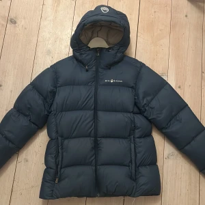 Blå dunjacka från Sail Racing - Säljer en blå dunjacka från Sail Racing i storlek large. Jackan har en dragkedja framtill och är långärmad med en mysig huva. Tyvärr har den en liten repa men går att  lämna in eller sy ihop själv. 