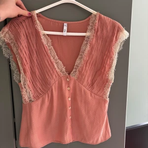 Rosa blus från Mango - Säljer en söt rosa blus från Mango med spetsdetaljer och knappar framtill. Blusen har en v-ringad design och är ärmlös med plisserade detaljer på axlarna. Perfekt för en somrig look!