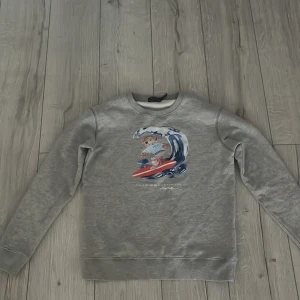 Grå sweatshirt från Ralph Lauren - Säljer en grå sweatshirt från Ralph Lauren med ett coolt tryck av en björn som surfar. Tröjan har långa ärmar och en rund halsringning. Perfekt för en avslappnad stil. 🐻🏄‍♂️