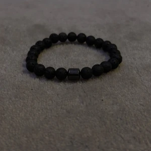 Svart lava pärlarmband - Snyggt svart armband med runda pärlor och en central metallisk detalj. Perfekt för att ge en stilren touch till din outfit.