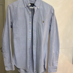 Blå skjorta från Polo Ralph Lauren - Snygg blå skjorta från Polo Ralph Lauren i slim fit. Skjortan har långa ärmar och knappar framtill samt en broderad logotyp på bröstet. Perfekt för en stilren look.
