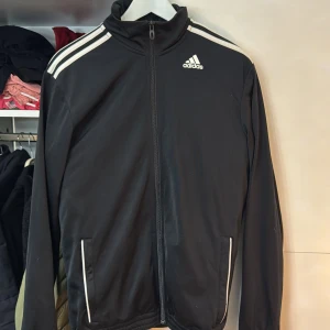 Svart träningsjacka från Adidas - Säljer en svart träningsjacka från Adidas med vita ränder på axlarna och dragkedja framtill. Jackan har två fickor med vita detaljer och Adidas-logga på bröstet. Perfekt för sportiga aktiviteter eller som en casual jacka. Storlek L men passar M för jag har det