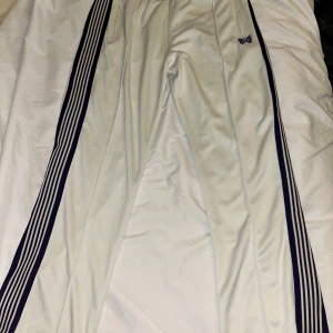 Needles track pants  - Säljer ett par vita träningsbyxor från Needles med lila fjärilsbroderi och lila ränder längs sidorna. Byxorna har elastisk midja och dragkedjefickor. Perfekta för en sportig look!