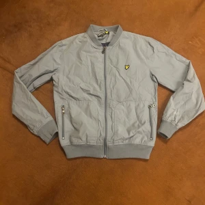 Blå bomberjacka från Lyle & Scott - Säljer en stilren grå bomberjacka från Lyle & Scott med dragkedja och ribbade muddar. Jackan har ett broderat emblem på bröstet och två praktiska fickor med dragkedja. Perfekt för en cool och avslappnad look.