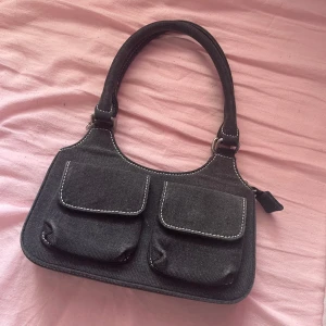 Svart axelväska från Brandy Melville  - Snygg svart axelväska i jeansmaterial med två praktiska fickor framtill. Väskan har en dragkedja upptill och en bekväm axelrem. Perfekt för att bära dina dagliga nödvändigheter med stil.