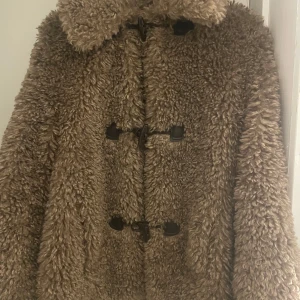 Brun teddyjacka med knäppning - Mysig brun teddyjacka med stora knappar framtill. Perfekt för kyliga dagar med sin fluffiga och varma design. Jackan har en klassisk krage och är långärmad.