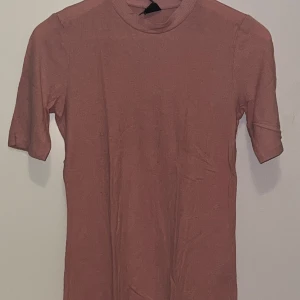 Rosa ribbad topp från Gina Tricot - En enkel och stilren rosa ribbad topp från Gina Tricot. Modellen har korta ärmar och en högre rund halsringning. Perfekt att matcha med jeans eller kjol för en avslappnad look.