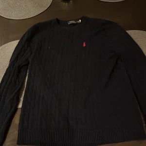 Mörkblå stickad tröja från Polo Ralph Lauren - Säljer en klassisk mörkblå stickad tröja från Polo Ralph Lauren med rund halsringning och röd broderad logga på bröstet. Tröjan har lång ärm och ribbade muddar vid ärmslut och nederkant. Perfekt för en stilren look. Priset är inte hugget i sten