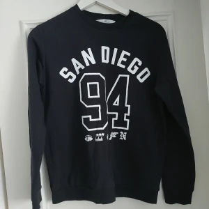 Svart sweatshirt med San Diego-tryck - Svart sweatshirt med vitt tryck 'San Diego 94' på bröstet. Tröjan har rund halsringning och långa ärmar. Perfekt för en avslappnad stil.