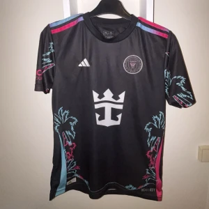Inter Miami CF svart fotbollströja Messi #10 - Snygg svart Inter Miami CF fotbollströja från Adidas med Messi #10 på ryggen. Tröjan har korta ärmar, tropiska palmmotiv i blått och rosa samt klubbmärke på bröstet. Perfekt för fans av laget och Messi.