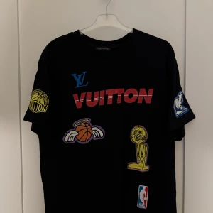 Svart Louis Vuitton x NBA t-shirt - Svart t-shirt från Louis Vuitton med NBA-tema och flera färgglada tryck, bland annat basketbollar, troféer och logotyper. Stort 'Vuitton' tryck på framsidan och 'Louis Vuitton' samt basketgrafik på baksidan. Perfekt för dig som gillar streetwear och basket. 