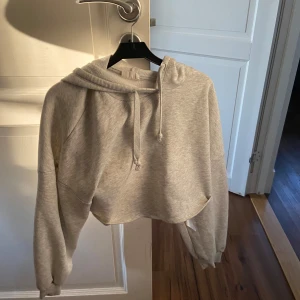 Ljusgrå croppad hoodie - Säljer en ljusgrå croppad hoodie med huva och dragsko. Tröjan har långa ärmar och är tillverkad i mjukt material, perfekt för en avslappnad stil. Passar dig som gillar en trendig och bekväm look.