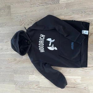 Svart hoodie från Hoodrich med duvtryck - Säljer en svart hoodie från Hoodrich med stort vitt tryck av duvor och text på både bröstet och ryggen. Hoodien har magficka och huva samt blå broderad text på ena ärmen. 