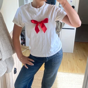 Vit t-shirt med röd rosett - Vit t-shirt med en stor röd rosett framtill. Klassisk rund halsringning och korta ärmar. Perfekt för dig som vill ha en enkel men ändå unik look.