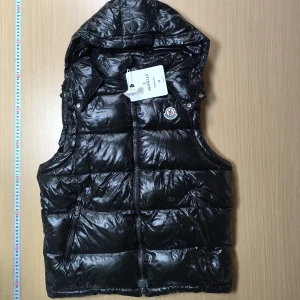 Svart dunväst med luva från Moncler - Säljer en svart, glansig dunväst från Moncler med luva och dragkedja framtill. Västen har två fickor med dragkedja och Moncler-logga på bröstet. Perfekt för lager-på-lager under kyliga dagar och det kommer med NFT SCAN