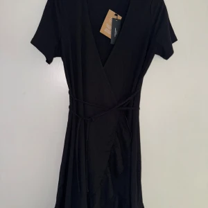 Svart omlottklänning från Vero Moda - Svart omlottklänning från Vero Moda med korta ärmar och knytband i midjan. Klänningen har en v-ringad hals och är tillverkad i ett lätt, strukturerat tyg. 