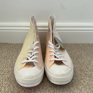 Converse Chuck Taylor All Star color changing sneakers - Säljer ett par Converse Chuck Taylor All Star high-top sneakers som skiftar färg. Skorna har en unik design med pastellfärger i beige, rosa och vitt, samt vit sula och tå. Klassisk logga på sidan och färgglad sula undertill. Dessa unika skor är vita från början men när de är utsatta i solen i några sekunder byter färgen
