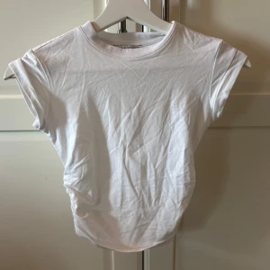 Vit croppad topp från Bershka - Säljer en vit croppad topp från Bershka med korta ärmar och rynkade sidor. Enkel och stilren design i mjuk bomull, perfekt till sommaren. Toppen har rund halsringning och en tight passform. Aldrig använt 