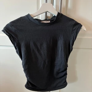 Svart croppad topp från Bershka - Säljer en svart croppad topp från Bershka i bomull med tight passform. Toppen har korta ärmar och rynkade detaljer i sidorna som ger en snygg effekt. Perfekt till sommaren och enkel att matcha med olika outfits. Använt få tals gånger. 