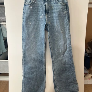 Blå bootcut jeans med blommiga detaljer från H&M - Säljer ett par blå bootcut jeans från H&M med baggy passform och justerbar midja. Jeansen har små blommiga detaljer i silver på framsidan som ger en unik look. Perfekta för dig som vill sticka ut lite extra.