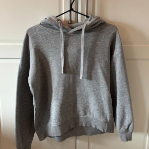 Grå hoodie från 157 - En enkel och stilren grå hoodie från lager157 med huva och dragsnören. Tröjan har långa ärmar och en klassisk passform, perfekt för en avslappnad look. Mjuk insida och ribbade muddar vid ärmslut och nederkant.