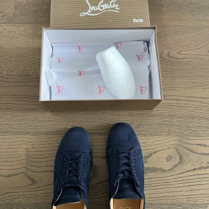 Mörkblå sneakers från Christian Louboutin - Snygga mörkblå sneakers från Christian Louboutin med klassisk röd sula och diskret broderad logga på hälen. Skorna har snörning och är tillverkade i mocka för en lyxig känsla. Perfekta för dig som vill sticka ut med stil.