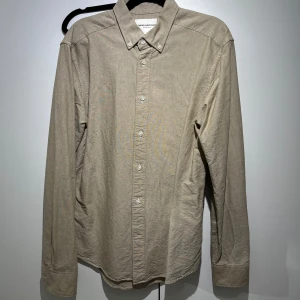 Beige skjorta - These Glory Days - Stilren beige skjorta från These Glory Days med klassisk krage och knäppning framtill. Skjortan har långa ärmar och en enkel, tidlös design som passar till många olika tillfällen.