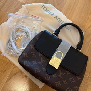 Handväska med svart lock - Säljer en exklusiv handväska med klassiskt brunt monogrammönster och svart lock. Väskan har guldfärgat lås, svart handtag och medföljer axelrem. Perfekt accessoar för dig som vill ha något lyxigt och tidlöst.