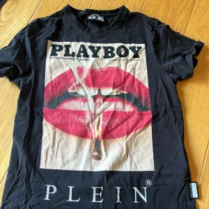 Svart t-shirt med Playboy-tryck från Philipp Plein - Cool svart t-shirt från Philipp Plein x Playboy med stort tryck av röda läppar och cigarett på framsidan. T-shirten har rund hals och är tillverkad i 100% bomull. Perfekt statement-plagg för dig som gillar unika designer.