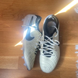 Nike fotbollsskor i vitt och svart - Säljer ett par Nike fotbollsskor i vitt med svarta detaljer och snörning. Skorna har rund tå, platt sula med dobbar och är tillverkade i syntetmaterial. Perfekta för dig som vill ha stilrena och funktionella skor på planen. Har även kvitto till reklamation 