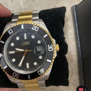Svart och guld herrklocka från GIW - Stilren herrklocka från GIW med svart urtavla, datumvisning och gulddetaljer. Armbandet är i tvåtonad metall, silver och guld. Klockan har tydliga indexmarkeringar och vridbar bezel. Extra länkar medföljer.
