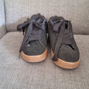 Svarta sneakers från DC Shoes - Säljer ett par svarta sneakers från DC Shoes med bruna gummisulor. Skorna har en enkel och stilren design med rund tå och DC-logga baktill. Perfekta för dig som gillar streetwear och skate-stil.