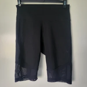Svarta cykelshorts från SOC - Svarta cykelshorts från SOC med hög midja och meshdetaljer längst ner på benen. Tillverkade i ett stretchigt syntetmaterial som andas och håller dig torr under träning. Perfekta för gymmet eller löprundan.