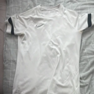 Vit Nike fotbollströja med svarta detaljer - Säljer en vit kortärmad fotbollströja från Nike med svarta detaljer på ärmarna och klassisk Nike-logga på bröstet. Perfekt för träning eller match. Materialet är lätt och andas bra.