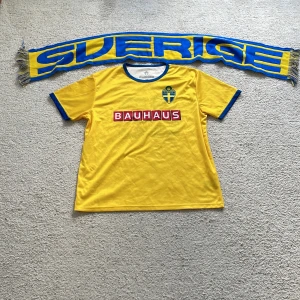 Sverige herrlandslagets fotbollströja gul/blå - F!ick den på landslags match.Gul fotbollströja med blå detaljer från Sveriges herrlandslag. Bauhaus-logga på bröstet och landslagets emblem på vänster sida. Rund hals med blå kant och blå ärmslut. Perfekt för att stötta Sverige på match