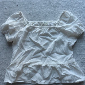 Vit blus med puffärmar från H&M - Säljer en söt vit blus från H&M med puffiga korta ärmar och fyrkantig ringning. Blusen har en volang nertill och är gjord i ett lätt, luftigt material. Perfekt för varma dagar och enkel att styla till olika outfits. Storleken är 170